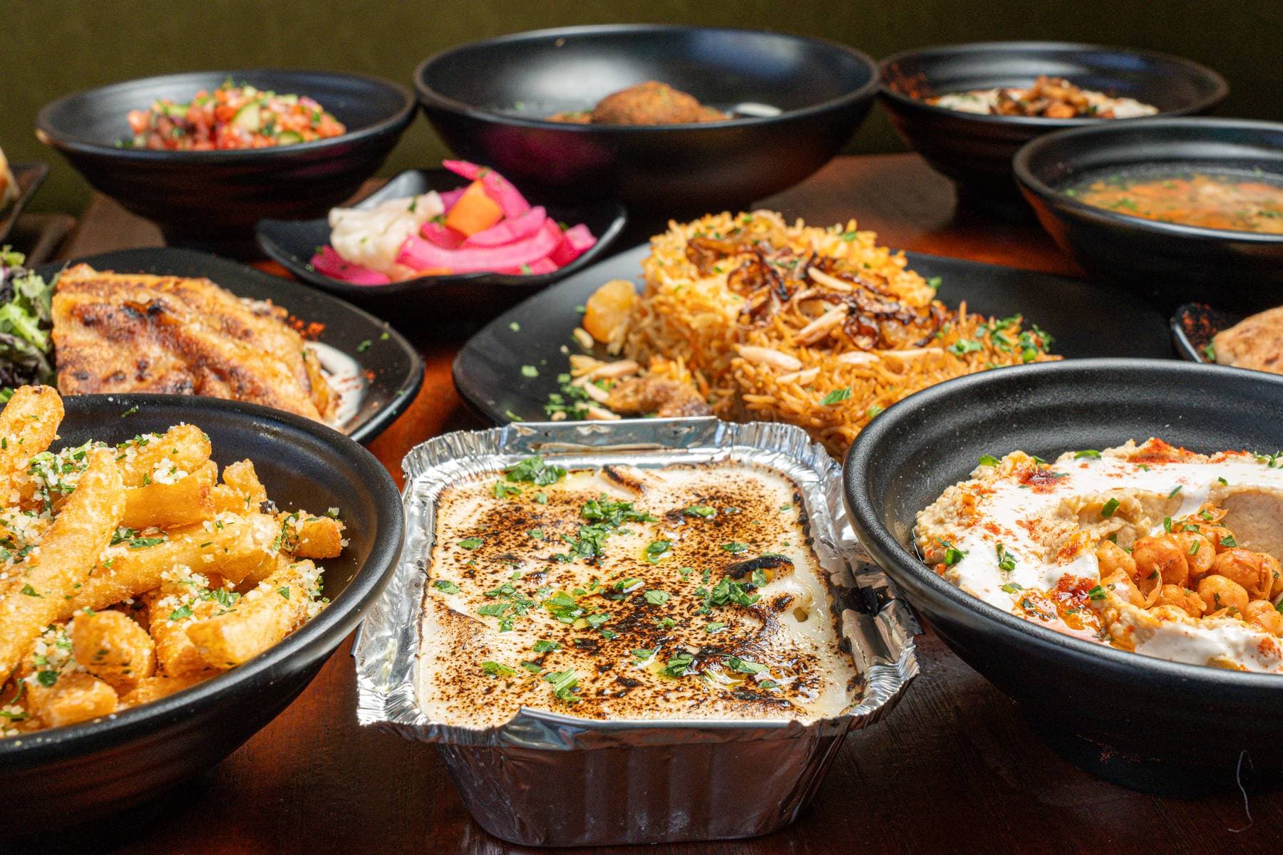 An overhead spread of small plates and bowls — hummus dusted with sumac, fries, salads, beans, and rice — laid out family-style for the table
